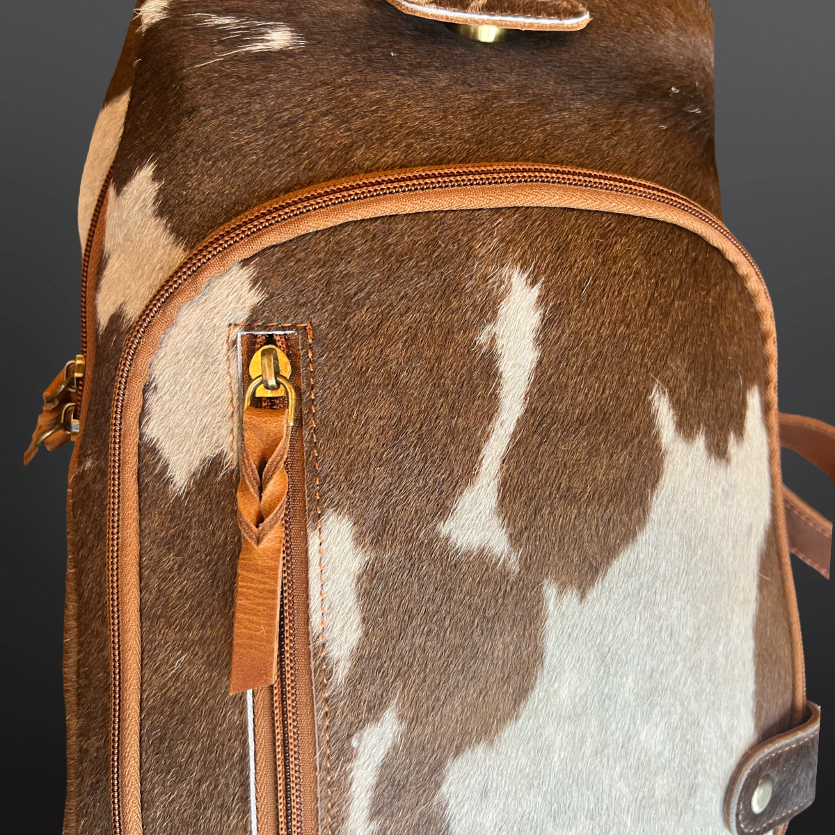 Brown and white cowhide leather bag with gold zipper pulls on a dark background