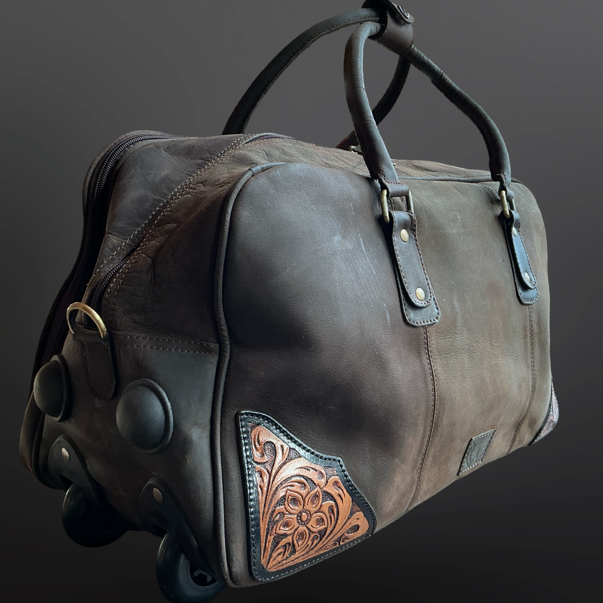 Brown leather duffel bag with tooled leather patches on a dark background